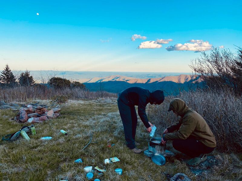 Two people are camping on a grassy hilltop with a scenic mountain view. One person in dark clothing is pouring something into a cooking pot, while another person in a tan jacket is crouched nearby. Camping gear and scattered items are on the ground, and the remnants of a campfire are visible to the left. The sky is blue with scattered clouds.